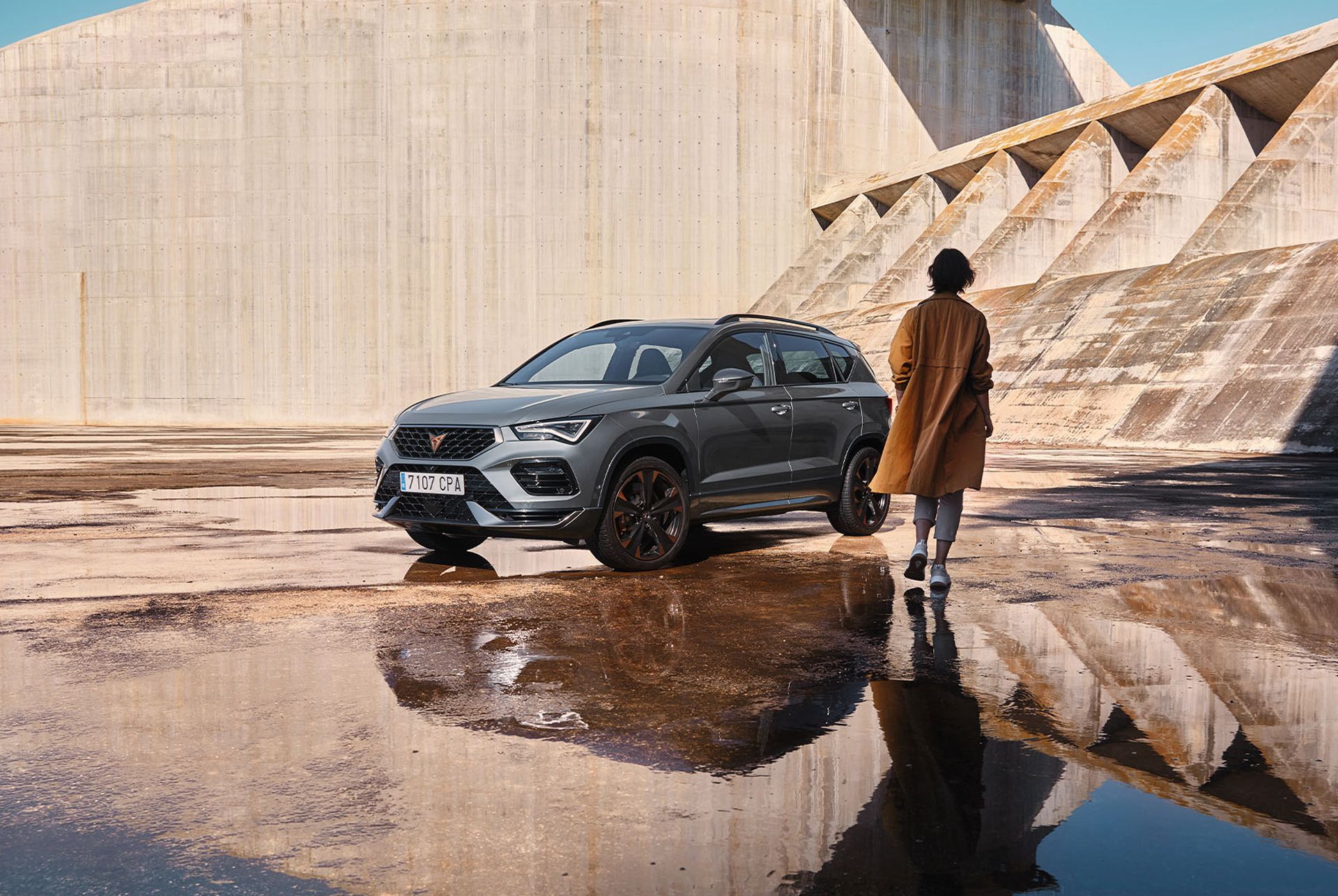 A CUPRA Ateca SUV in Graphite Grey, featuring copper alloy wheels, parked in a concrete area with a model approaching.