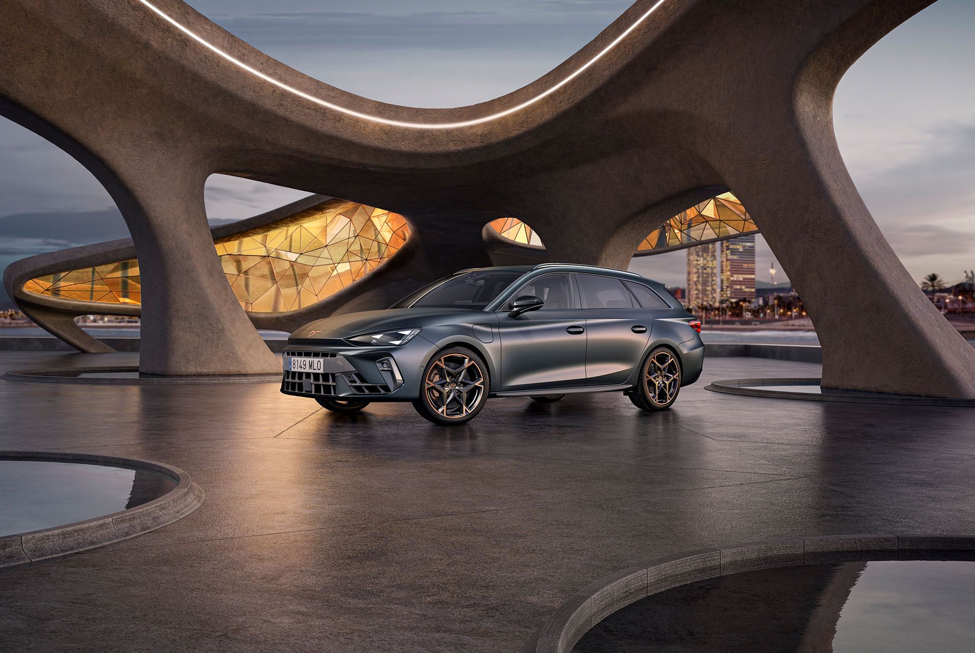 A three quarter front view of the CUPRA Leon Sportstourer in enceladus grey matte with copper accent alloy wheels parked on smooth concrete under an architectural structure. Barcelona cityscape in the background.