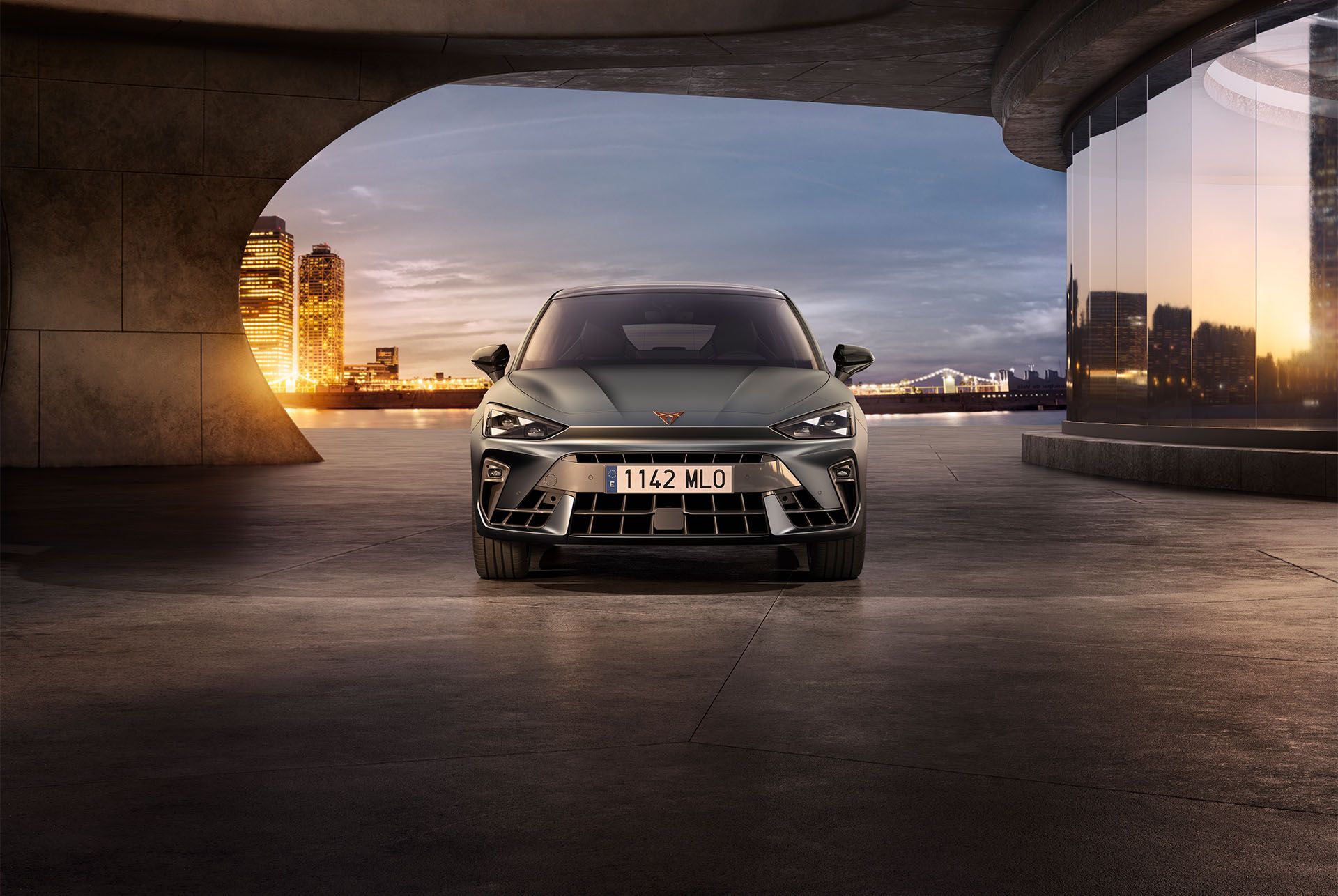 A front facing view of the CUPRA Leon in enceladus grey matte with copper accent alloy wheels parked on smooth concrete under an architectural structure. Barcelona cityscape in the background.