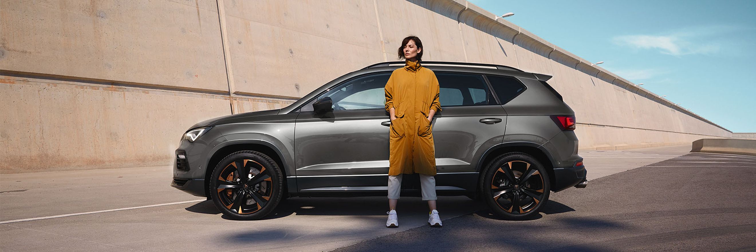 A passenger side view of the CUPRA Ateca SUV in graphite grey with a model standing on front of the vehicle.