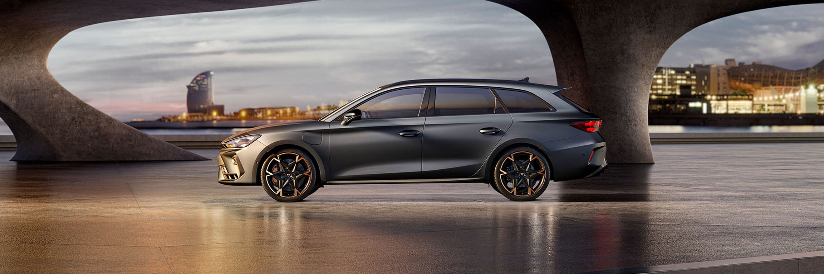 A side view of the CUPRA Leon Sportstourer in enceladus grey matte with copper accent alloy wheels parked on smooth concrete under an architectural structure. Barcelona cityscape in the background.