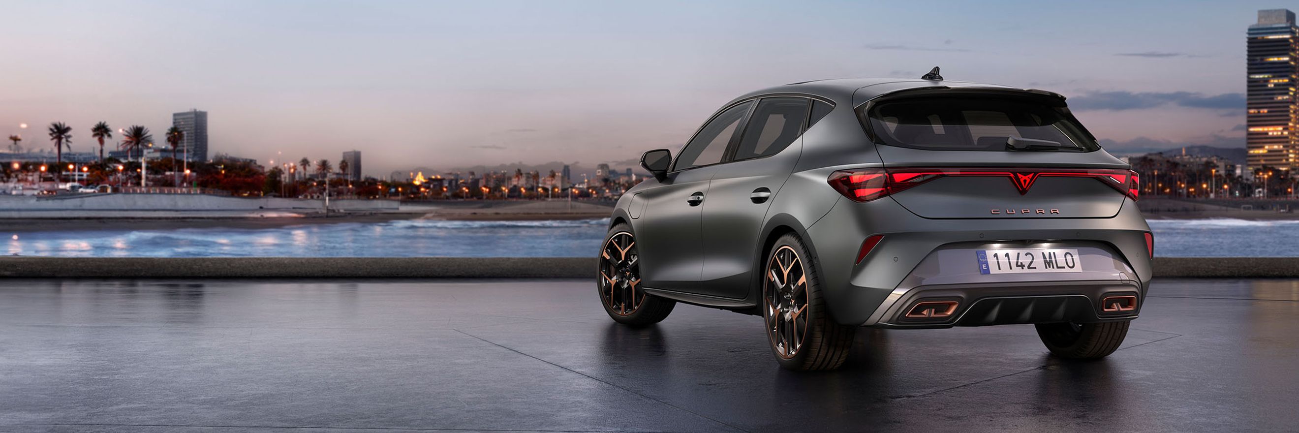 A rear three quarter view of the CUPRA Leon in enceladus grey matte with copper accent alloy wheels parked on smooth concrete with the barcelona cityscape in the background.